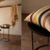 Cojín de lino con rayas en colores tierra y azul y una manta a juego colgada en una barra de madera con pared blanca de fondo.