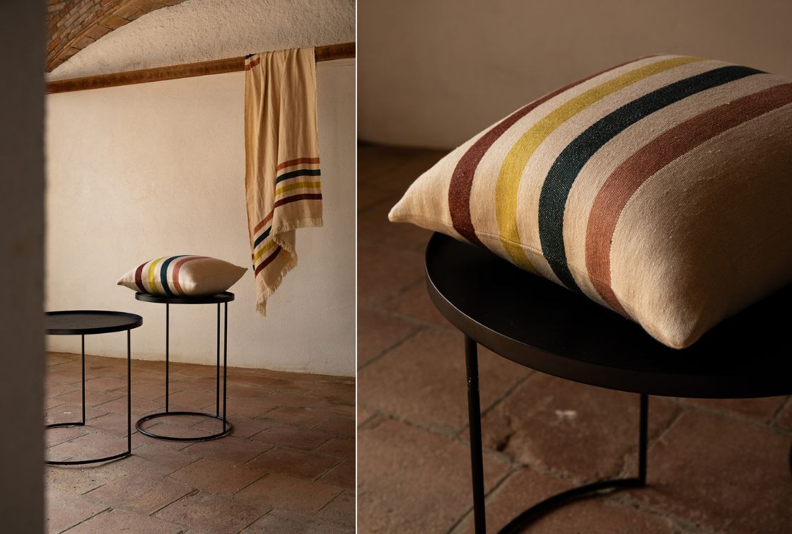 Cojín de lino con rayas en colores tierra y azul y una manta a juego colgada en una barra de madera con pared blanca de fondo.
