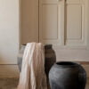 Dos jarrones de cerámica negra, uno con una manta de lino beige colgada, colocados junto a una pared blanca y una puerta de madera blanca