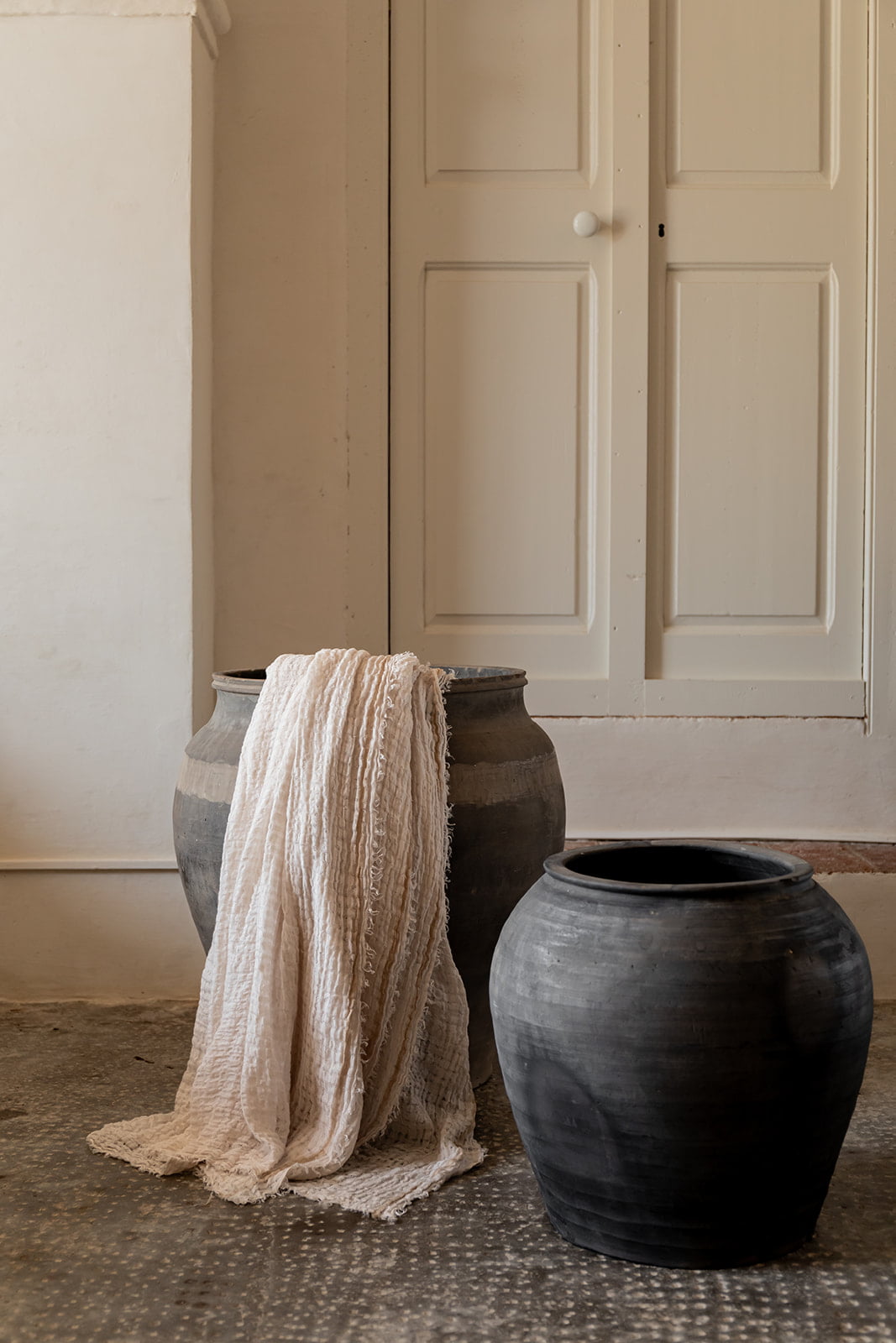 Dos jarrones de cerámica negra, uno con una manta de lino beige colgada, colocados junto a una pared blanca y una puerta de madera blanca