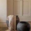 Dos jarrones de cerámica negra, uno con una manta de lino beige colgada, colocados junto a una pared blanca y una puerta de madera blanca