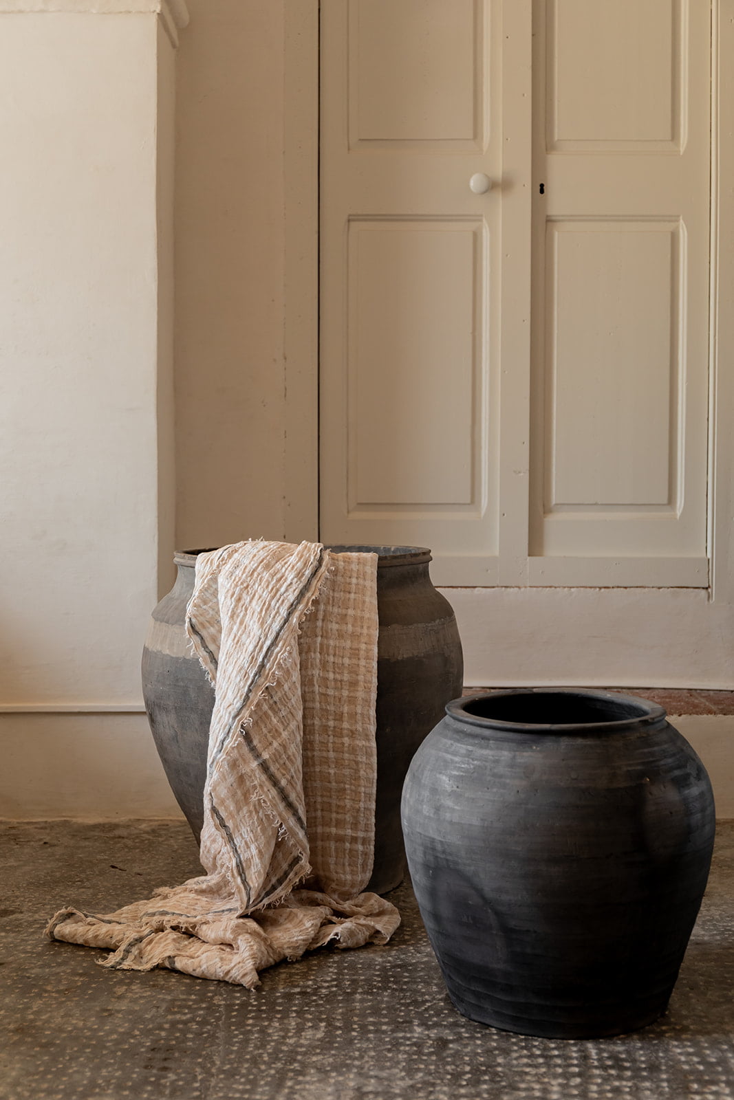 Dos jarrones de cerámica negra, uno con una manta de lino beige colgada, colocados junto a una pared blanca y una puerta de madera blanca