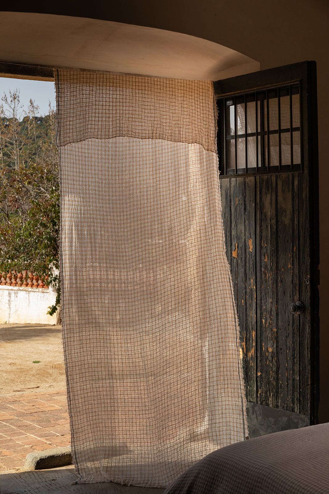 Cortina de lino beige y marrón colgada al lado de una puerta de madera desgastada con una ventana con rejas