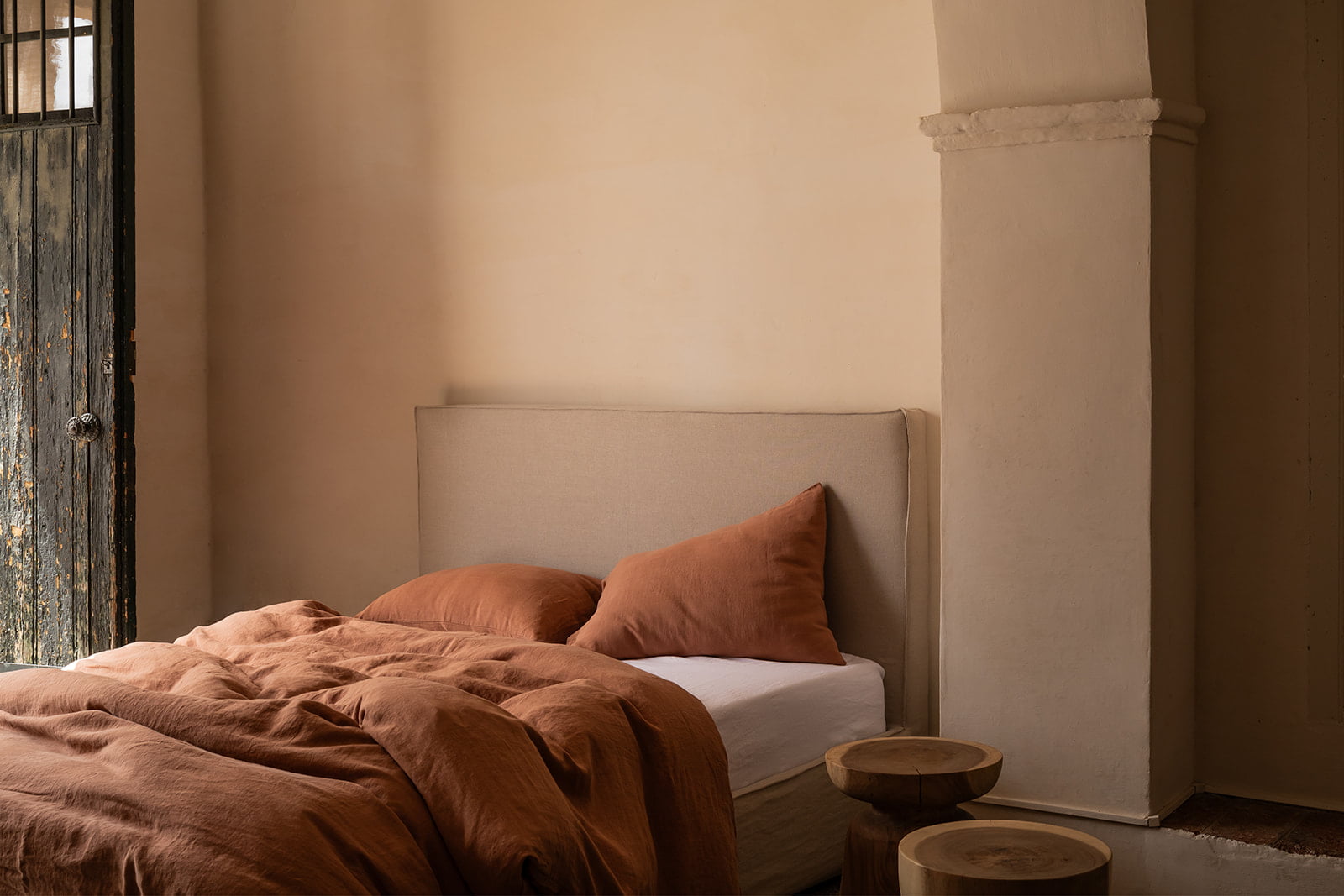 Cama con cabecero beige y ropa de cama en tonos marrones, acompañada de un taburete de madera