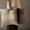 Cojín de lino beige con rayas negras, sostenido por una persona de pie.