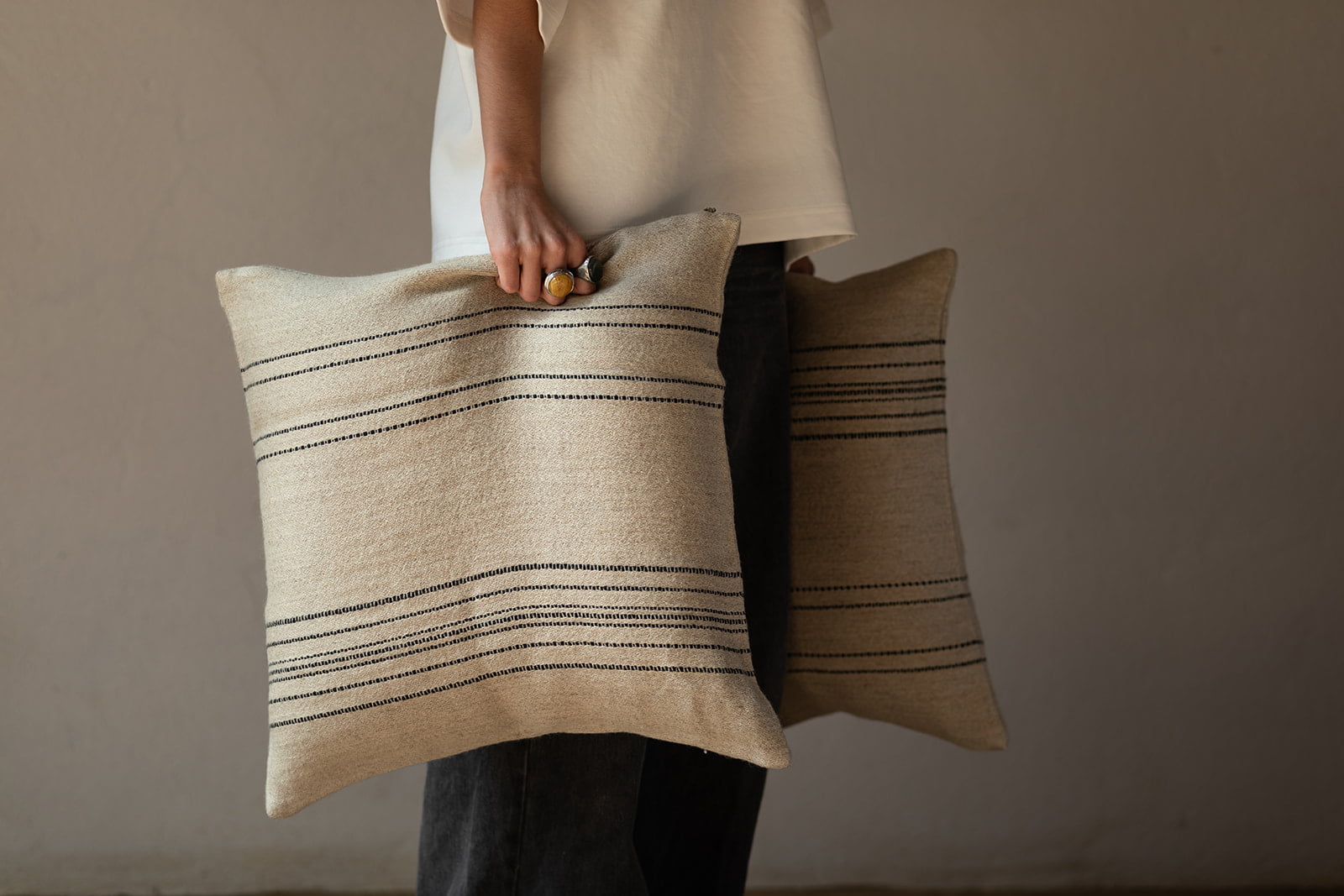 Cojín de lino beige con rayas negras, sostenido por una persona de pie.