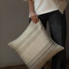 Cojín de lino beige con rayas negras, sostenido por una persona de pie.