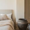 Cama con cabecero beige y funda nórdica de lino en color blanco, con un jarrón de barro grande al lado en un dormitorio minimalista.