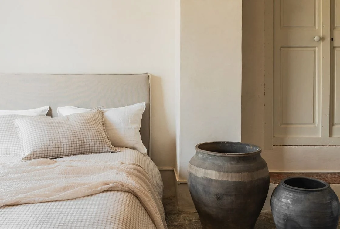 Cama con cabecero beige y funda nórdica de lino en color blanco, con un jarrón de barro grande al lado en un dormitorio minimalista.