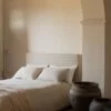 Cama con cabecero beige y funda nórdica de lino en color blanco, con un jarrón de barro al lado en un dormitorio iluminado.
