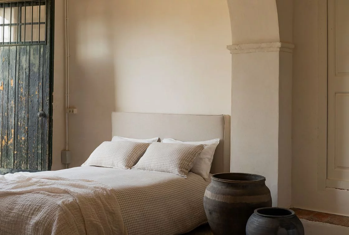Cama con cabecero beige y funda nórdica de lino en color blanco, con un jarrón de barro al lado en un dormitorio iluminado.