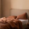 Cama con cabecero beige y funda nórdica de lino en color canela, acompañada de cojines a juego en un dormitorio minimalista.