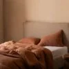 Cama con cabecero beige y funda nórdica de lino en color canela, acompañada de cojines a juego en un dormitorio minimalista.