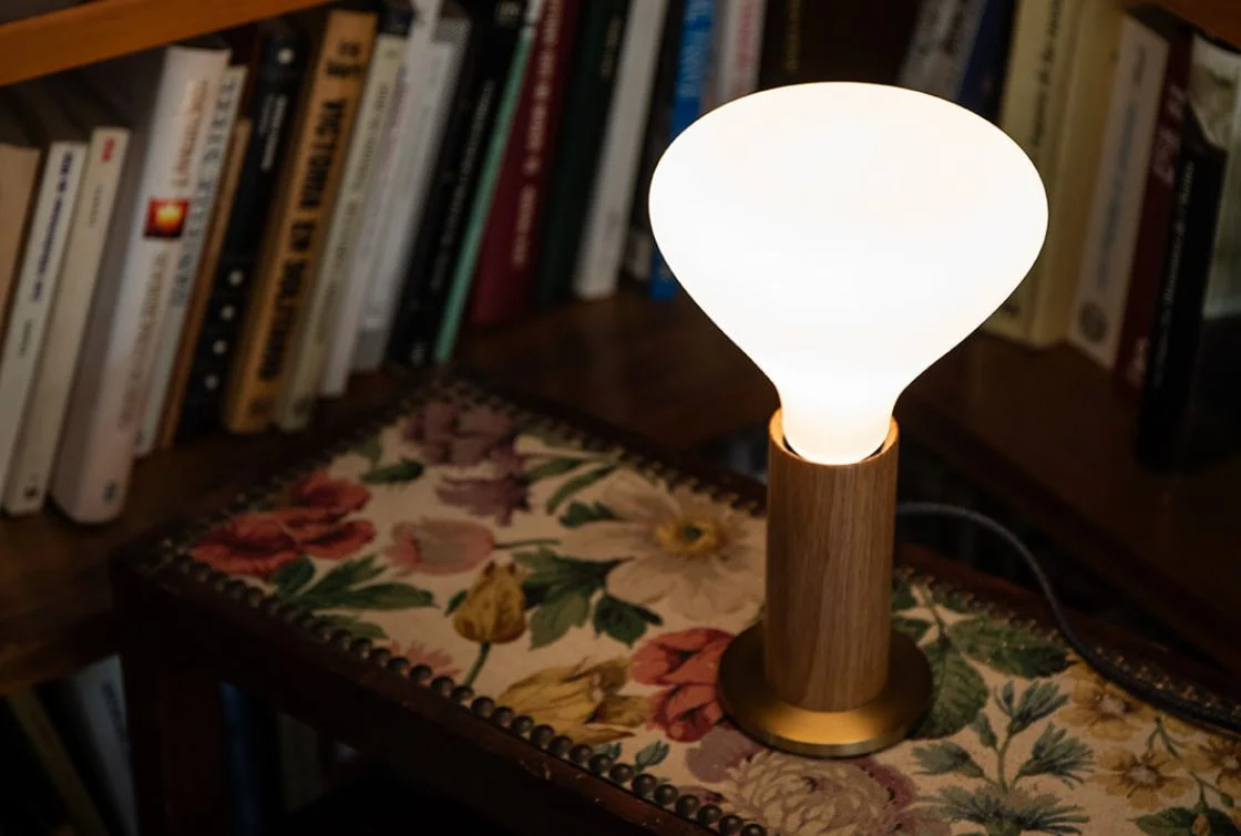 Lámpara de mesa con base de madera y pantalla blanca, colocada sobre una mesa junto a libros
